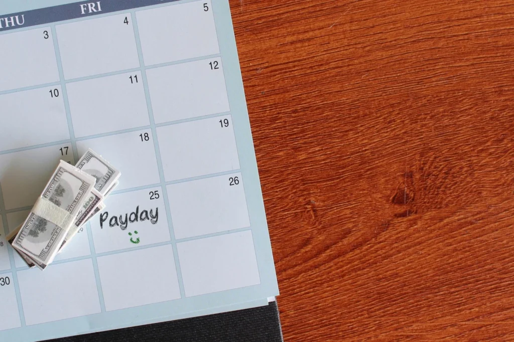 Calendario mensual apoyado sobre un escritorio de madera, con un fajo de billetes de cien dólares sobre los casilleros y la palabra Payday escrita a mano en uno de los días