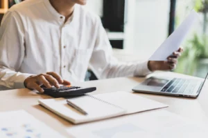 Analista de nómina usa una calculadora mientras revisa un documento impreso, con una notebook y un cuaderno sobre el escritorio