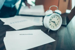 Sobre de papel y planillas impresas sobre un escritorio de madera, junto a un reloj despertador blanco, mientras una persona revisa documentos de fondo