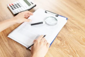 Manos revisando con lupa y lapicera una planilla de liquidación con columnas de importes sobre un escritorio de madera, junto a una calculadora